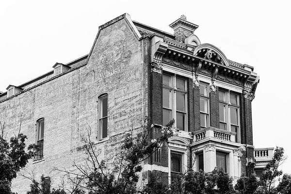 Black and white photograph of the historic Reuter Building on Alamo Plaza in San Antonio, Texas. In addition to it's impressive front facade, the brick side displays a beautiful multi-layered expanse of faded vintage ads. The Reuter Building is a four-story brick building with carved stone located on the corner of Crockett Street and Alamo Plaza. It was designed James Wahrenberger for William Reuter in 1891, and was operated as a saloon and ladies’ parlor.