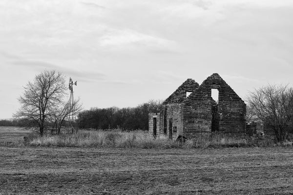 Black and white fine art photograph of an abandoned broken stone farmhouse with a windmill on the American prairie