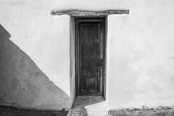 Black and white architectural photograph of a wooden door in the thick adobe wall of the old Spanish Mission San Juan Capistrano, which was established in San Antonio by Franciscans in 1731. The church was completed in 1756.