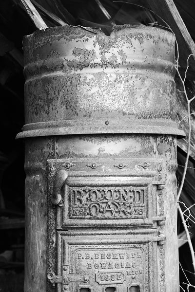Black and white fine art photograph of a rusty antique Round Oak stove discovered in a collapsing building in a small town. The words on the face of the stove read "Round Oak No. 18, 1895, P.D. Beckwith, Dowagiac, Mich".