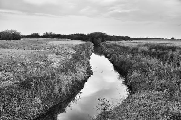 Black and white fine art photograph of a glassy creek winding through the undulant American prairie landscape