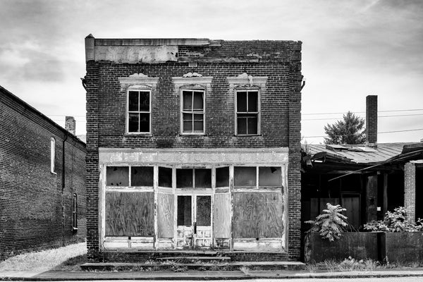 Black and white photograph of an empty storefront on the abandoned Main Street in the ghost town of Pamplin City, Virginia. The street is lined with 11 historic, vacant buildings.