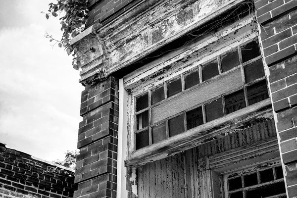 Black and white photograph of frames of colored glass over the front door of an abandoned building in a small town. The portico is covered with cracked white paint. Over the door is a faded sign that says "Estate."
