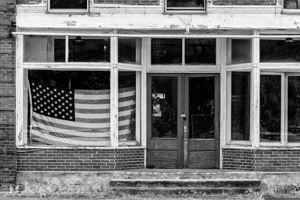 Black and white photograph of an abandoned storefront with a US flag in the window on the deserted old Main Street in Pamplin City, Virginia.