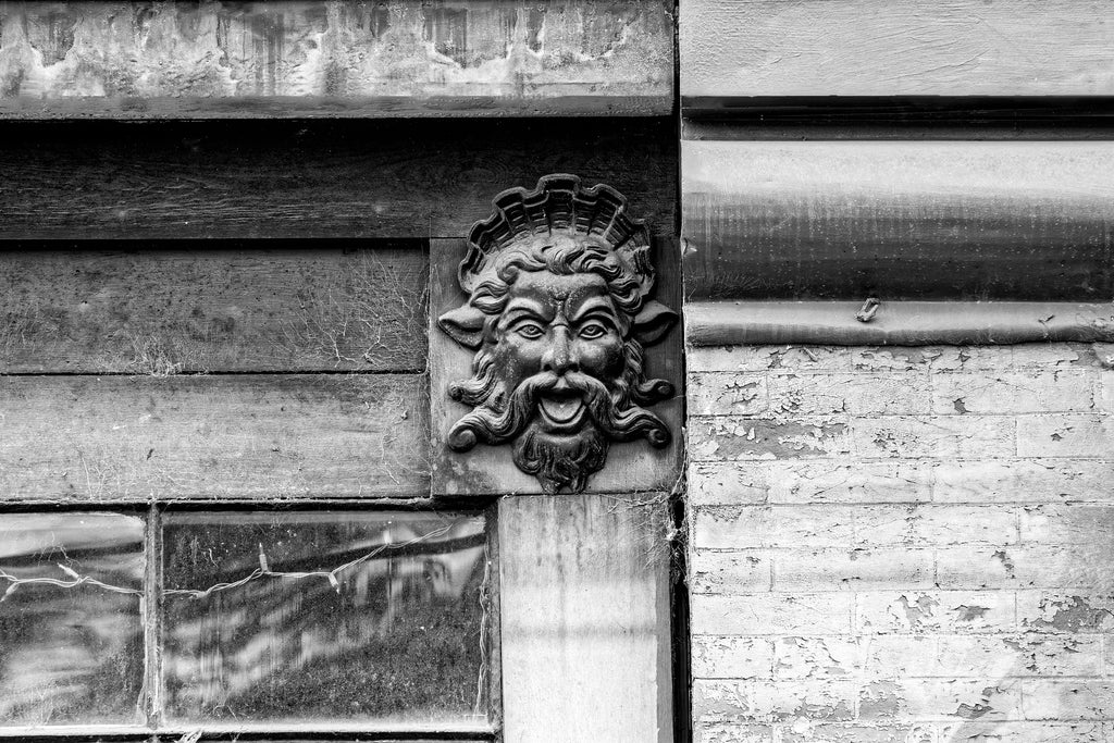 Black and white architectural detail photograph of the dusty head of a Greek god on the outside of a shabby old building.