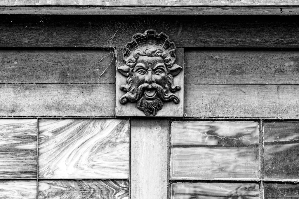 Black and white architectural detail photograph of a dusty gargoyle head looking reminiscent of the Greek god Poseidon.