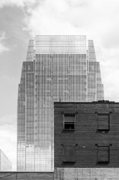 Black and white architectural photograph of Nashville's 2010 glass skyscraper called The Pinnacle at Symphony Place, towering over an old brick building probably dating to the 1890s-1900s. The Pinnacle at Symphony Place is 29 stories, 417 feet tall, and on sunny days like shown here, seems to become a mirage in the sky.