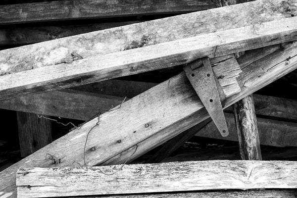 Black and white photograph of angled beams and a rusty metal hinge found in the shadows of a fallen old hay barn.