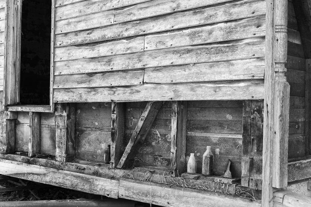 Black and white photograph of old bottles lined up on the side of an abandoned old farmhouse.