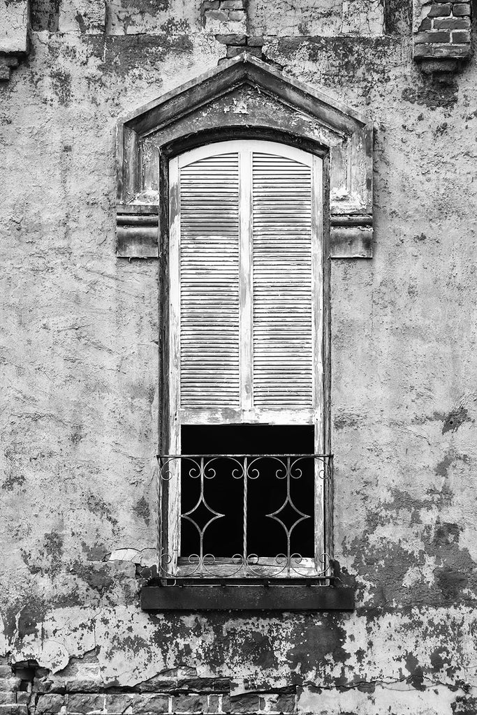 Black and white photograph of the second story window of an abandoned building with peeling paint, chipped bricks, and white shutters.