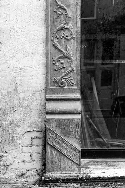 Black and white architectural detail photograph of an Mesker Iron Works metal column and nameplate on an abandoned building. Mesker Iron Works of Evansville, Indiana was one of the nation's leading sellers of iron work storefronts, tin ceilings, awnings, and other metal architecture elements. By 1915, they had sold storefronts into every state.