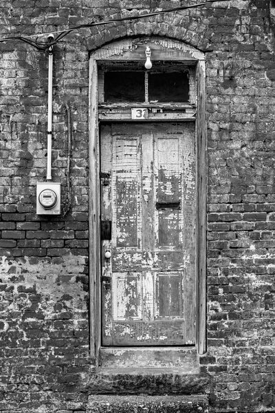 Black and white architectural detail photograph of a textured, old wooden door with chipped paint, found in an alley in a small town. Set into a brick wall with fading paint, this door has loads of detail and character.