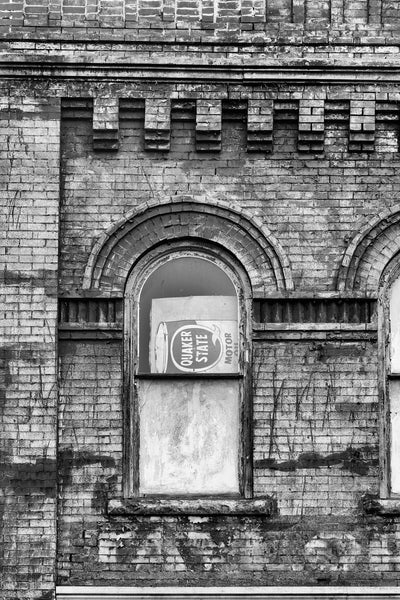 Black and white photograph of an arched second floor storefront window with a sideways motor oil sign and faded paint that says "Taylor" on dark bricks. 