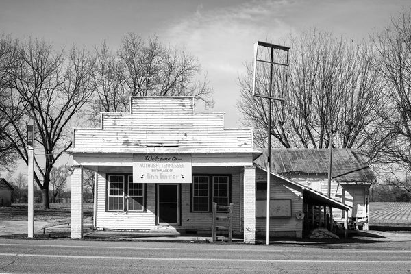 Black and white architectural photograph of an abandoned old clapboard storefront on Highway 19 in Nutbush, Tennessee. An old sign on the building says, "Welcome to Nutbush, Tennessee, Birthplace of Tina Turner." Farm fields can be seen behind the building.