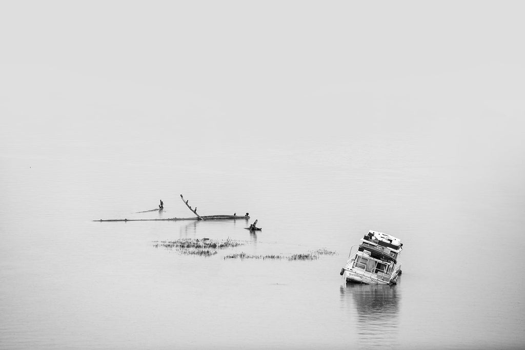 Black and white photograph of a boat sitting off-kilter after being run aground in the shallow part of a wide river.