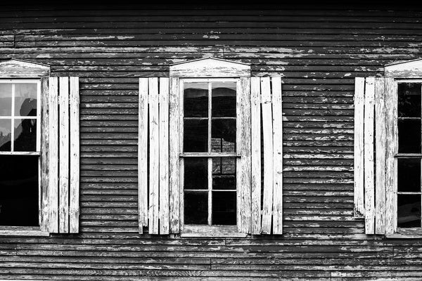 Black and white architectural detail photograph of rustic white shutters on the sides of an old red clapboard house.