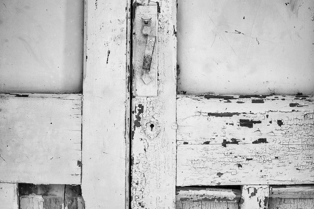 Black and white photograph of details in cracked paint and a bent ornate door handle on the doors of an abandoned building in a small town.