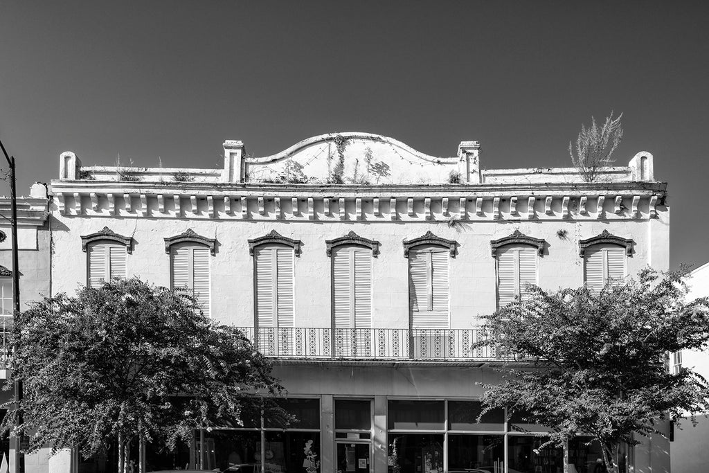 Black and white photograph of a historic storefront in downtown Natchez, Mississippi with a traditional ironwork balcony and plants growing from the roofline.