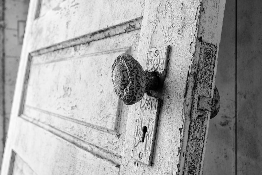 Black and white photograph of an ornate door knob on a detached door leaning against the wall inside an abandoned farm house.