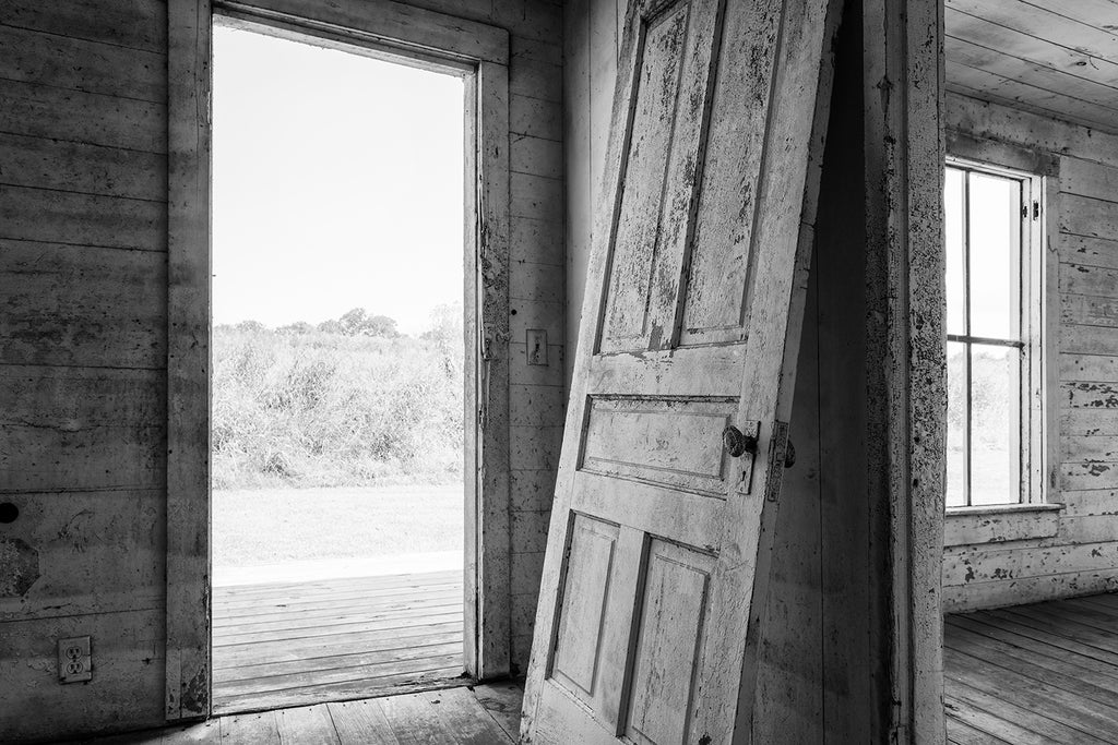 Black and white photograph of an old detached door leaning against the wall inside an abandoned farm house with fields visible outside.