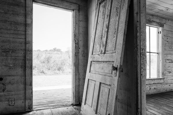Black and white photograph of an old detached door leaning against the wall inside an abandoned farm house with fields visible outside.