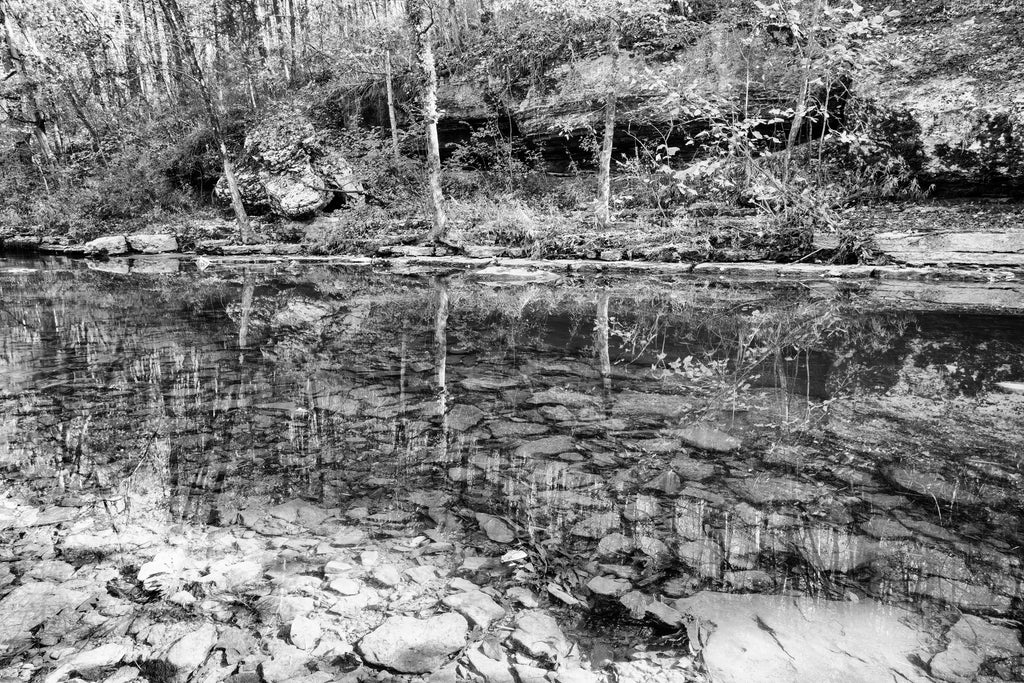 Black and white photograph of a placid stream with reflections of the landscape on its glassy surface and rocks visible below the surface