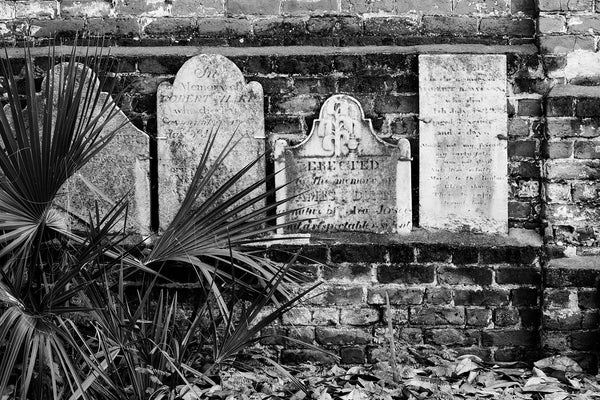 Black and white photograph of a row of headstones on a brick wall that borders Savannah's historic Colonial Cemetery. Established in 1750, the cemetery quickly expanded to six acres and was closed to interments in 1853. It now serves as a public park in Savannah's historic district.