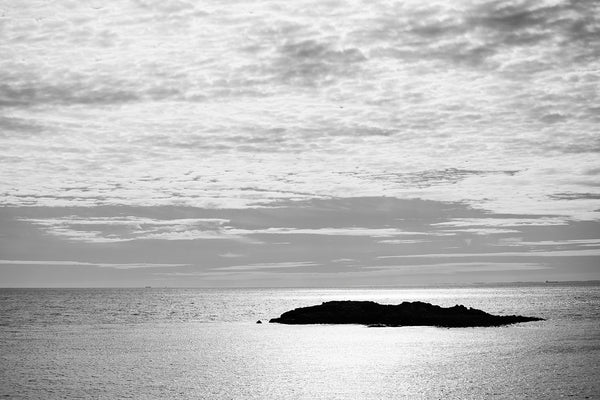 Black and white photograph of a small coastal island silhouetted in the bright light of sunrise off the New England shore.