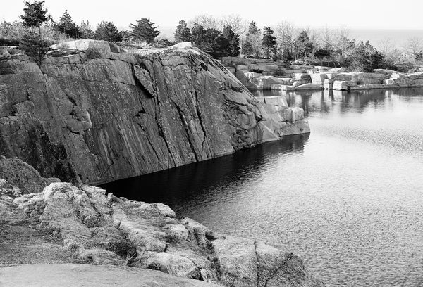 Black and white landscape photograph of a New England pond in a former granite quarry with a view of the Atlantic ocean in the distance.