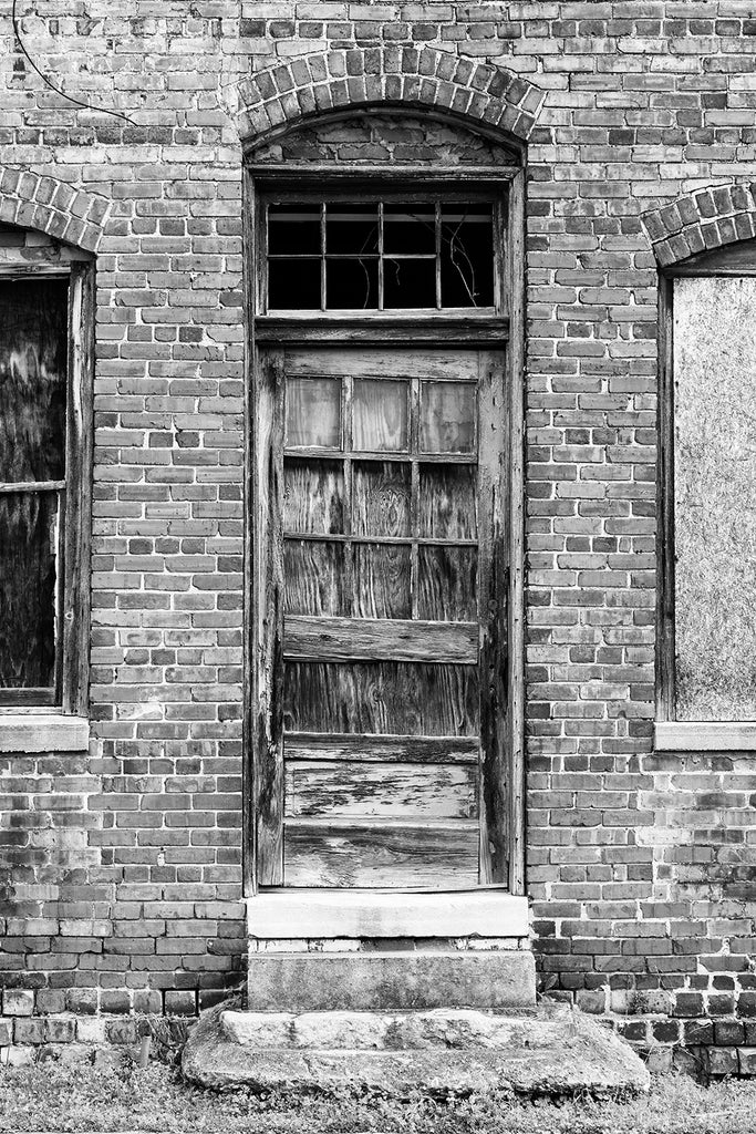 Black and white photograph of a weathered old wooden door on the exterior of an abandoned red brick mill building.