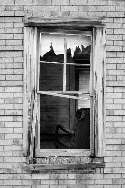 Black and white photograph looking of a window to an abandoned old house with a tattered chair and other left-behind items inside.