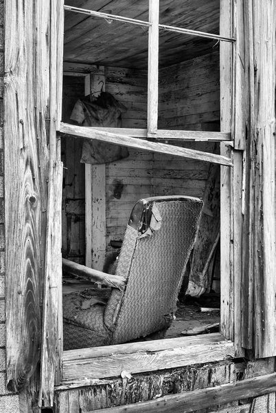 Black and white photograph looking into the window of an abandoned old house with a tattered chair and other left-behind items inside.