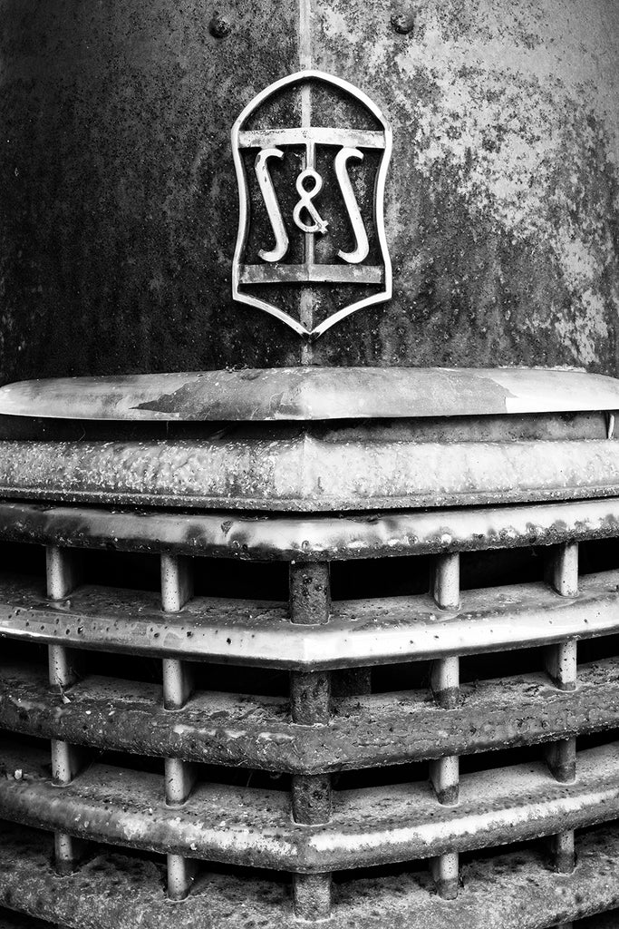 Black and white detail photograph of a rusty antique Sayers and Scovill automobile grill with a chrome hood ornament that says S&S.