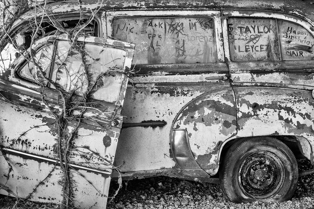 Black and white photograph of a rusty antique car covered with poison ivy and graffiti scribbled into the dust on the windows.