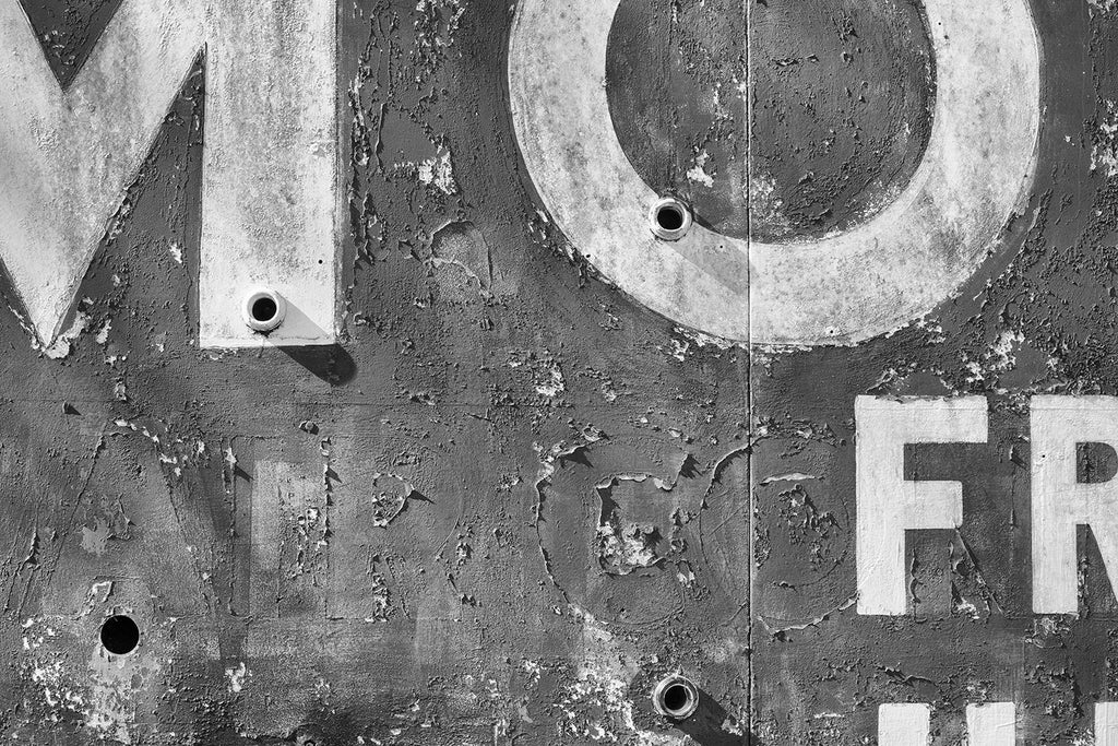 Black and white detail photograph of a textured old motel sign with peeling paint and ghost letters.