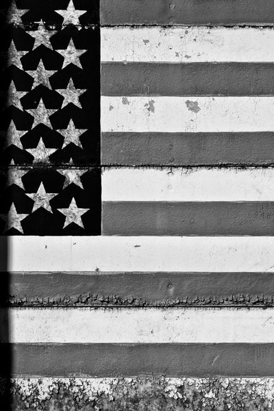 Black and white photograph of a rustic US flag painted on a factory door with seams running through it, and weathered decay along the edges.
