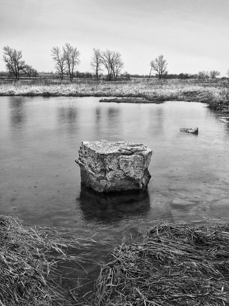 Black and white landscape photograph of a rock in the middle of the Pipestone Creek on the chilly Minnesota prairie in early Spring.