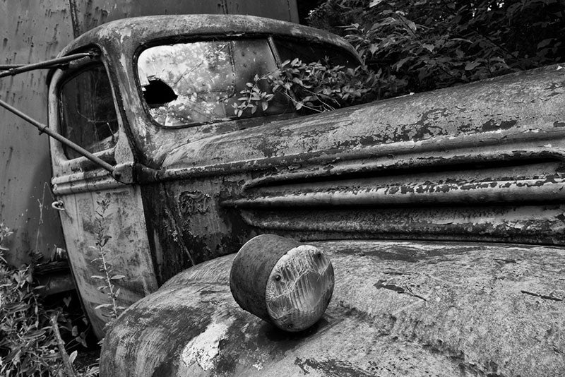Black and white fine art photograph of a wrecked and abandoned delivery truck. Someone drove this hand-painted antique truck, into a ditch on a winding country road, and never returned to retrieve it. It still sits there rusting away among the tall weeds and grass.