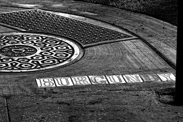 Black and white fine art photograph of the Burgundy street sign letters in the sidewalk beside a manhole cover in the French Quarter of New Orleans, Louisiana. 