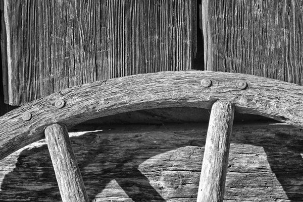 Black and white detail photograph of an old wooden wagon wheel leaning against a textured wooden wall.