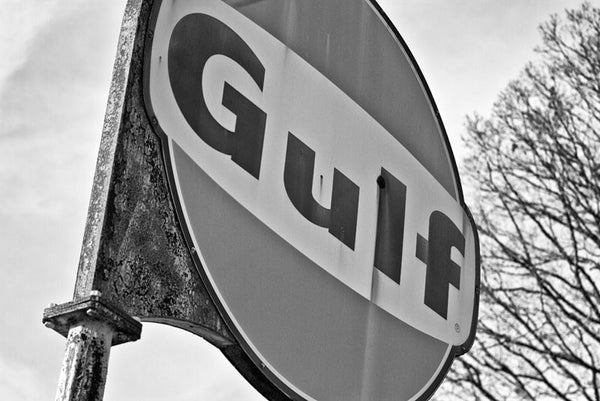 Black and white photograph of a rusty and fading Gulf gas station sign that still stands outside a long-vacated and abandoned service station on a rural highway in the southern US.