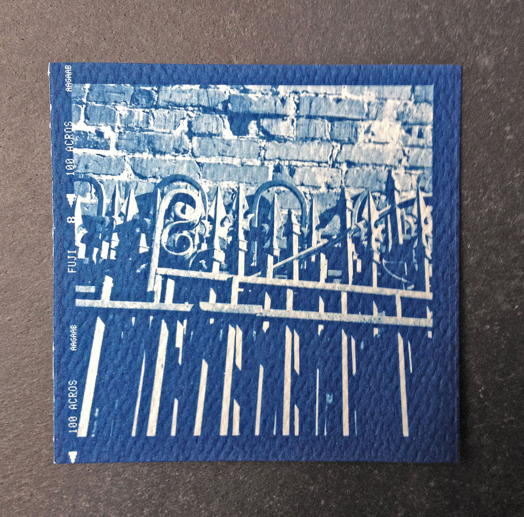 This is a unique, small, one-of-a-kind handmade cyanotype print of a many sets of antique ironwork fences stacked against a brick wall. This was contact printed from a 2-1/4 inch black and white film negative on textured ivory watercolor paper, handmade and printed by the artist.