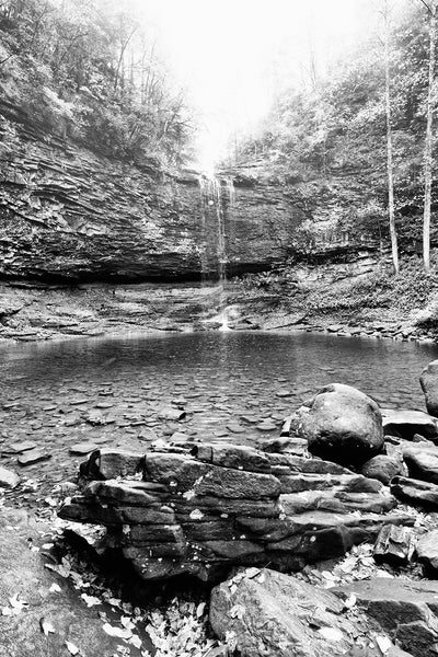 Black and white landscape photograph of the rocky pool in the basin below a tall mountain waterfall.