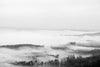 Black and white landscape photograph of dense morning fog passing over and between the trees and rolling peaks of an eastern mountain valley.