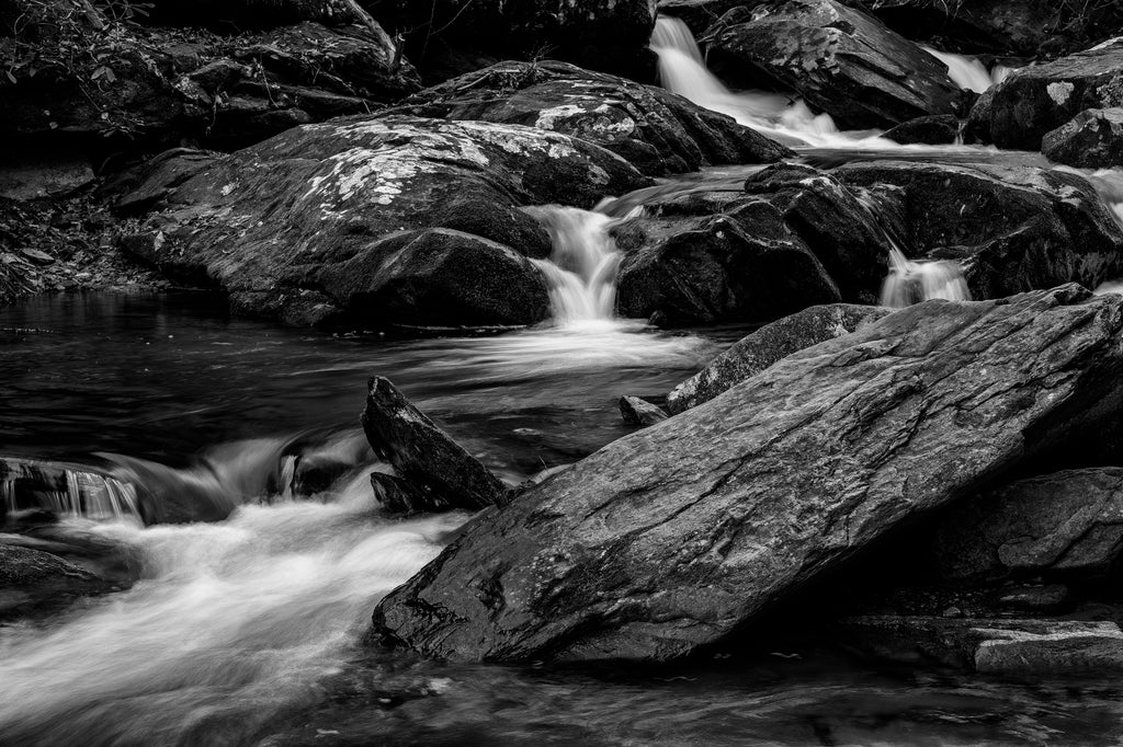 Black and white landscape photograph of a mountain stream pouring over and between large rocks and boulders on its way down hill.