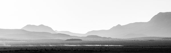 Black and white panoramic landscape photograph that shows a pickup truck crossing the desert at sunrise, leaving a cloud of dust in its wake, with layers of rugged western mountains in the background.