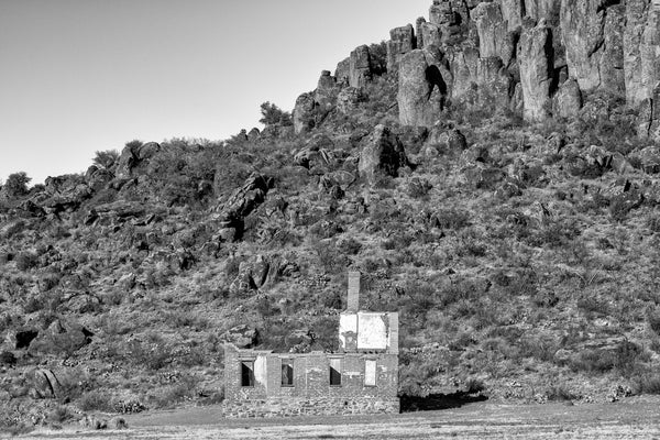 Black and white photograph of a big abandoned brick ruin at the base of a mountain in the western desert.