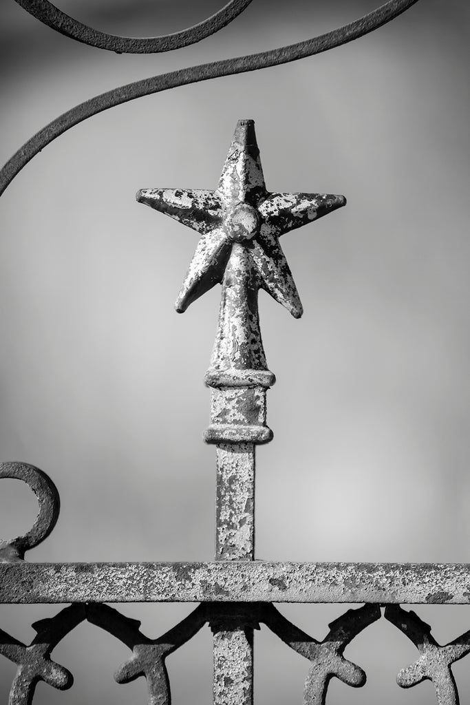 Black and white photograph of a tarnished ironwork star finial on an old fence surrounding an abandoned old house in far West Texas.