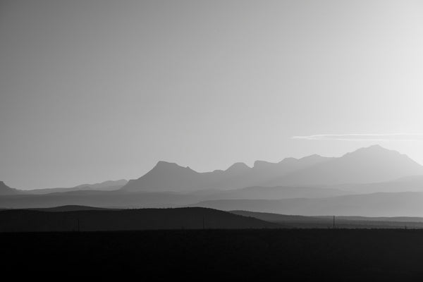 Black and white landscape photograph of a hazy mountain sunrise casting light across layers of ridges and peaks.