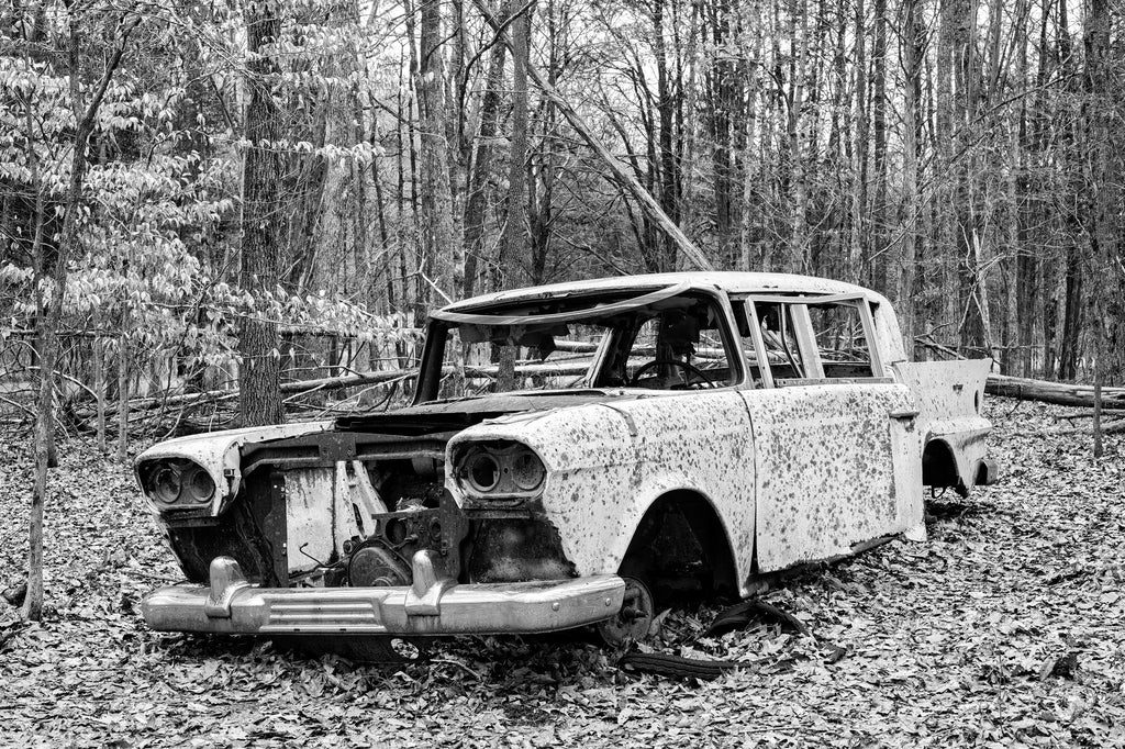 Black and white photograph of an abandoned classic automobile left sitting in the forest.